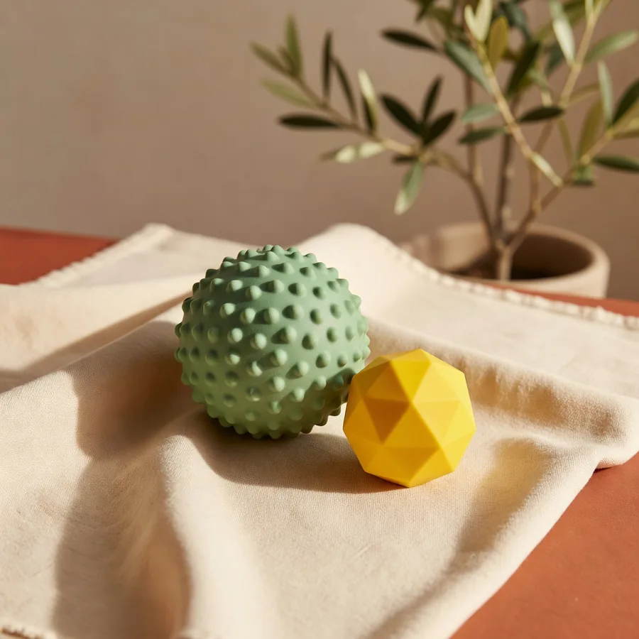 A sage TPR treat ball beside a smaller yellow faceted TPR ball on a cream sampling cloth